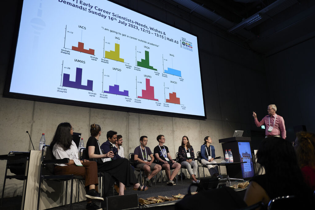 8 Early Career panellists are seated onstage in front of a screen of histograms at the IUGG23 'Big Theme' event. The Secretary General of IUGG stands at a lectern with a microphone.